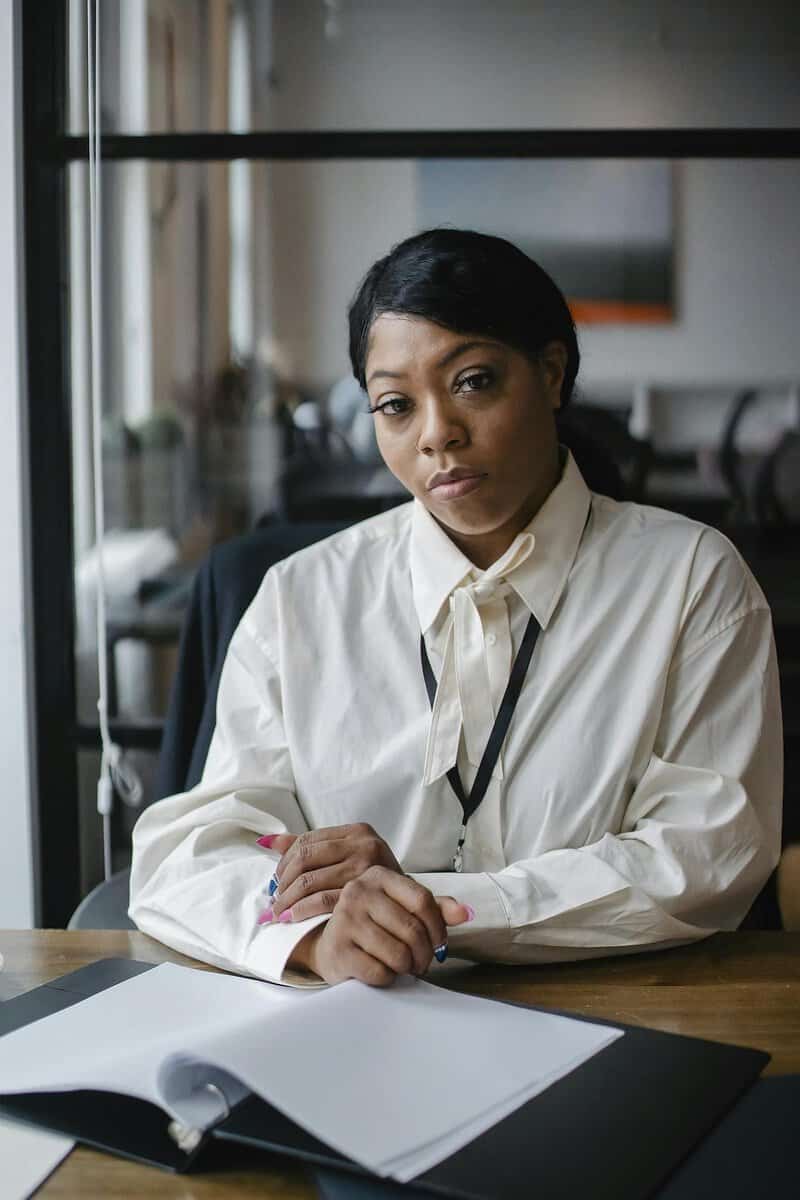 A woman in a white blouse sits at a desk with an open notebook, looking directly at the camera in an office setting.