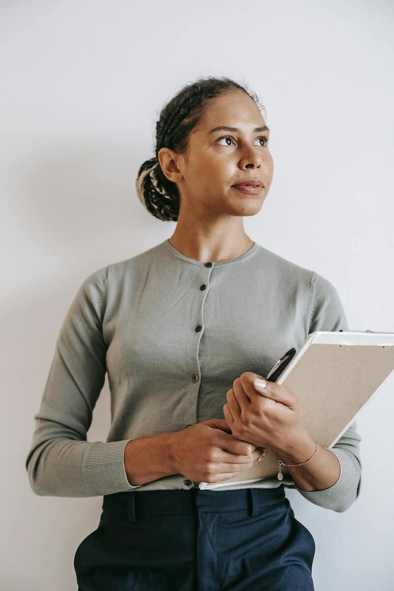 A woman in a light green cardigan holds a clipboard and pen while looking to the side against a plain white background.