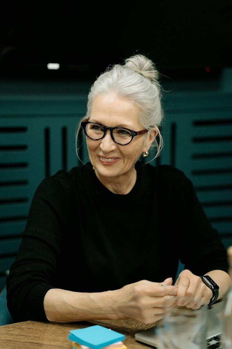 An older woman with gray hair in a bun, wearing glasses and a black top, sits at a table holding a pen with notepads and a laptop nearby.